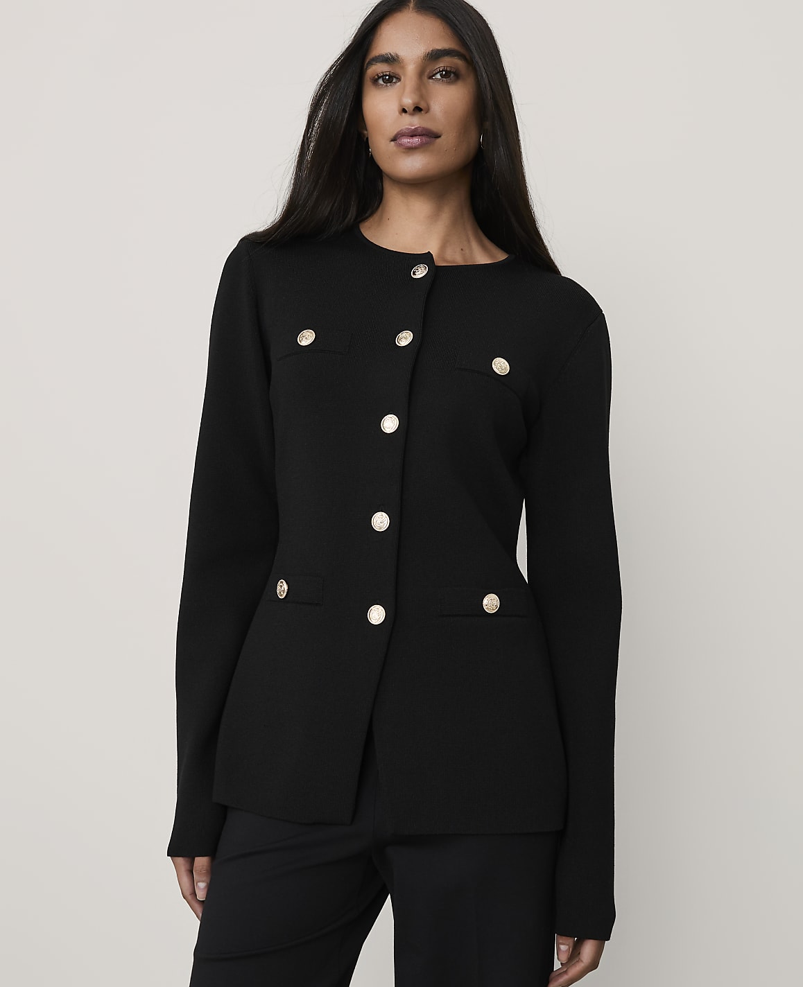 A woman wearing a black buttoned blazer and matching trousers stands against a plain, light-colored background.