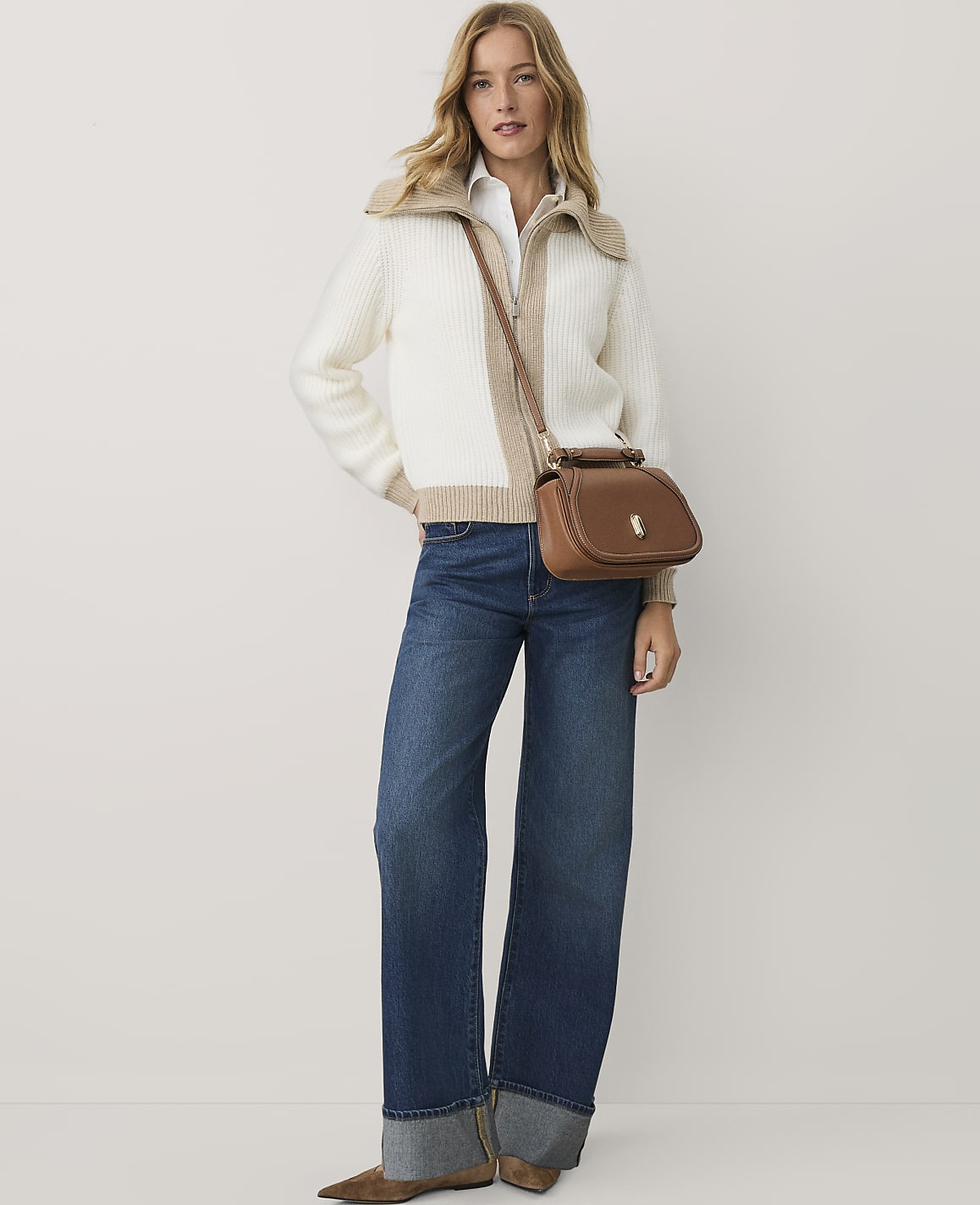 A woman wearing a white and beige zip-up sweater, blue wide-leg jeans, and a brown crossbody bag stands against a plain white background.