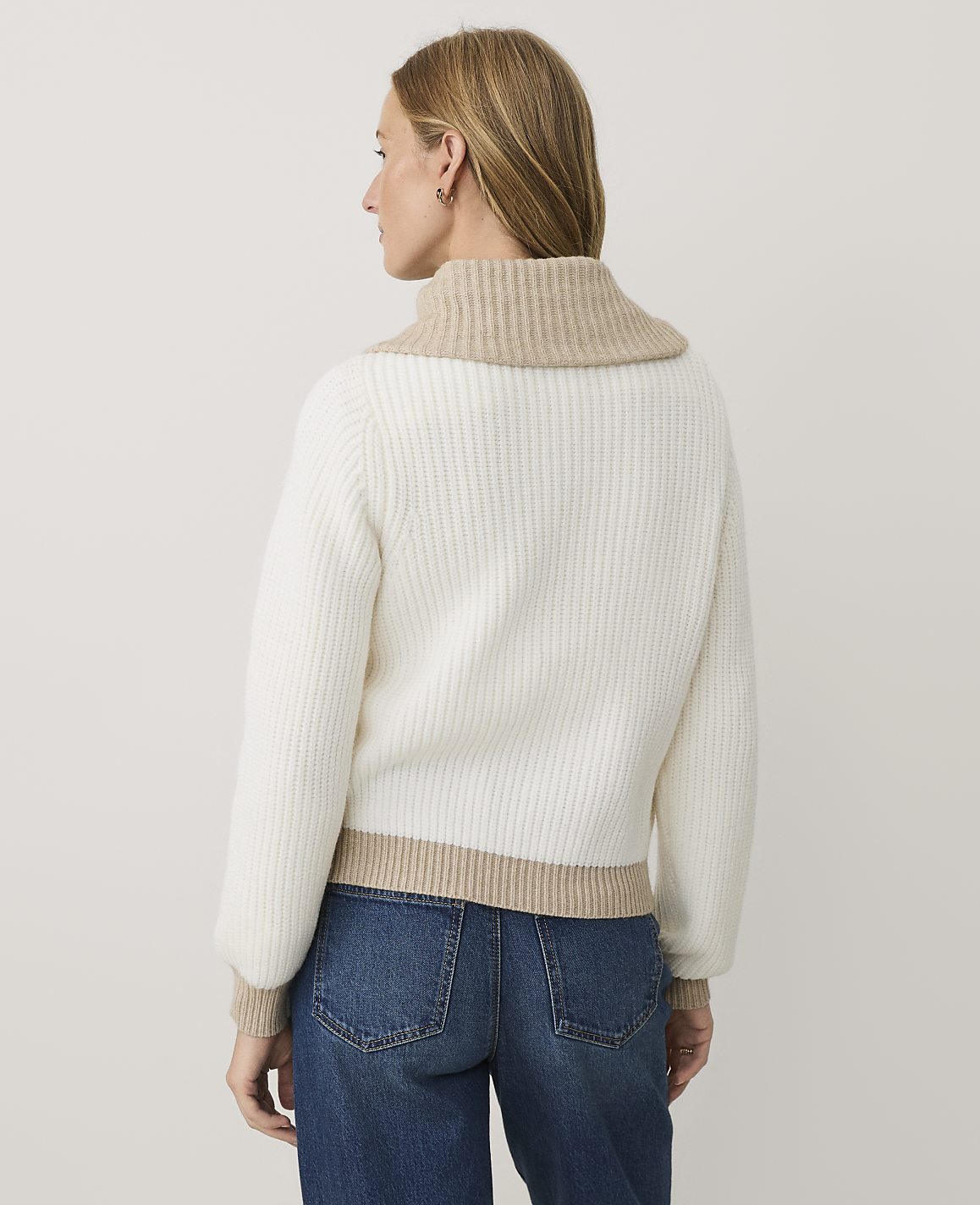 A woman wearing a white ribbed sweater with beige trim and blue jeans stands against a plain white background.