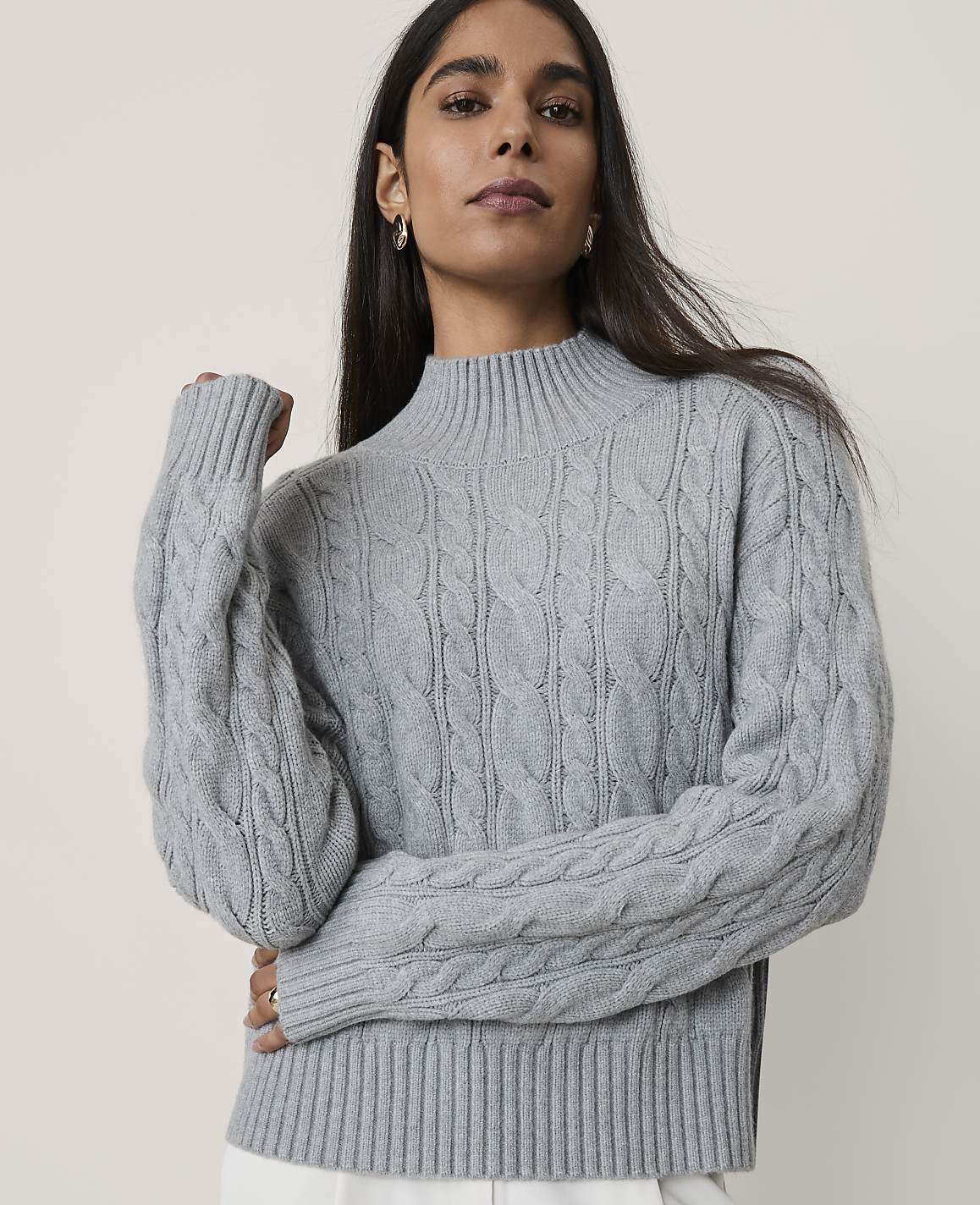 A woman wearing a grey cable-knit sweater stands against a plain, light-colored background.