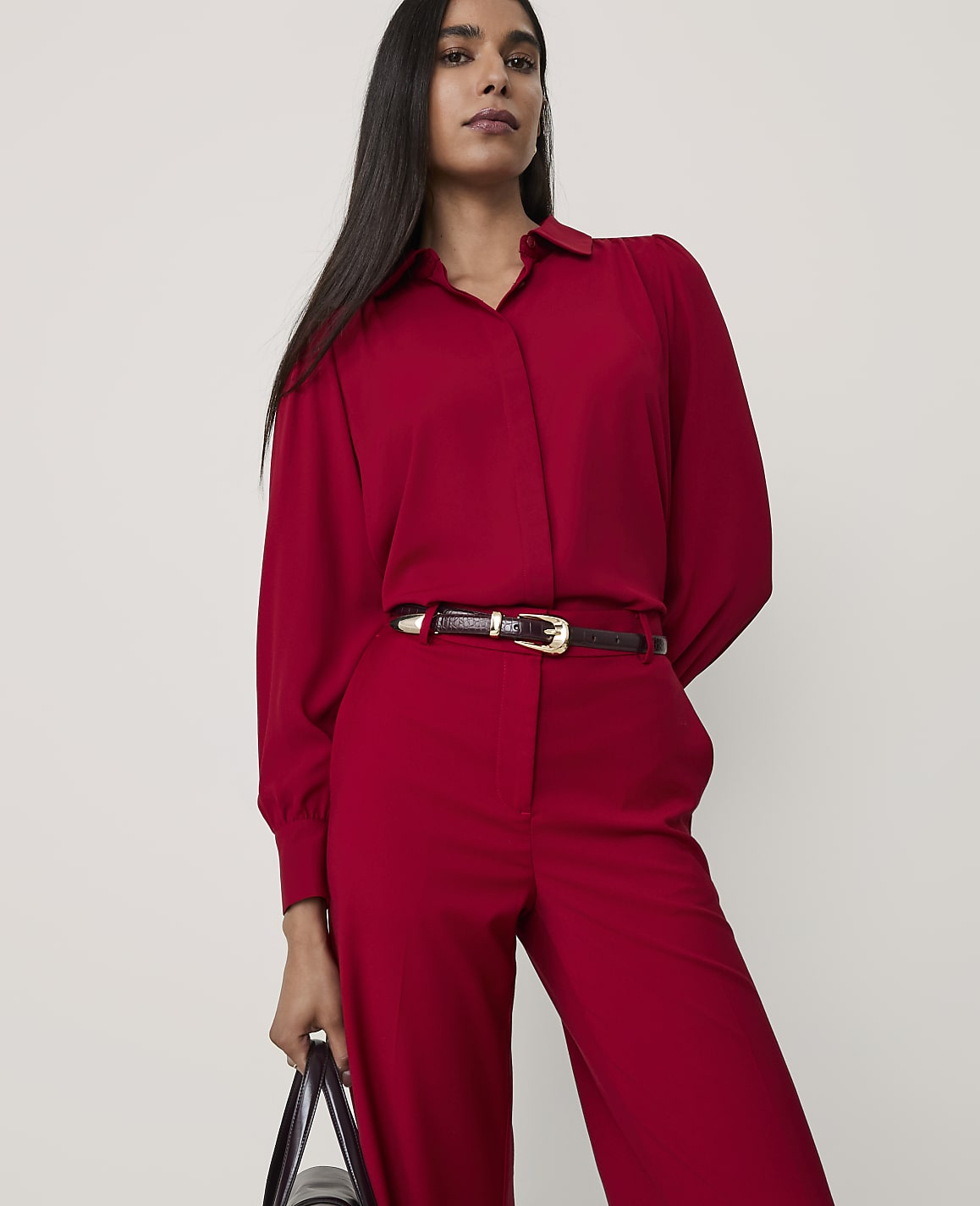 A woman wearing a red button-down shirt and matching trousers with a dark belt stands against a plain, light-colored background.