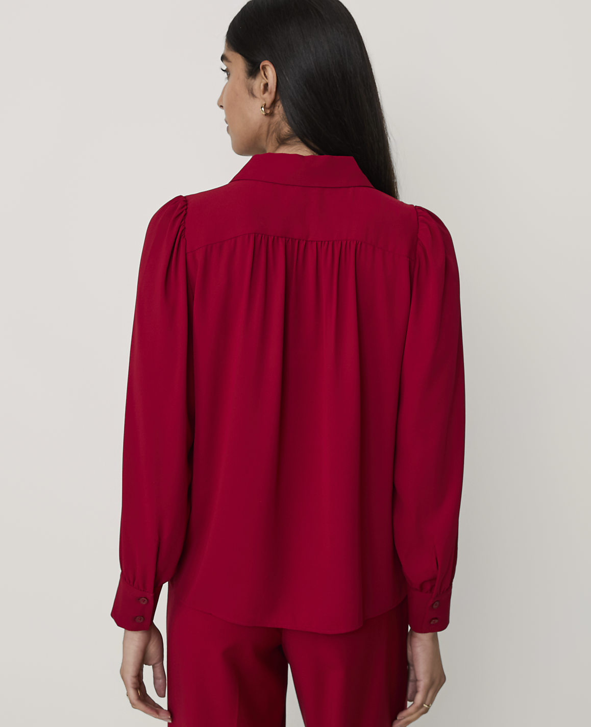 A woman wearing a red long-sleeved blouse and matching trousers stands with her back to the camera against a plain light gray background.