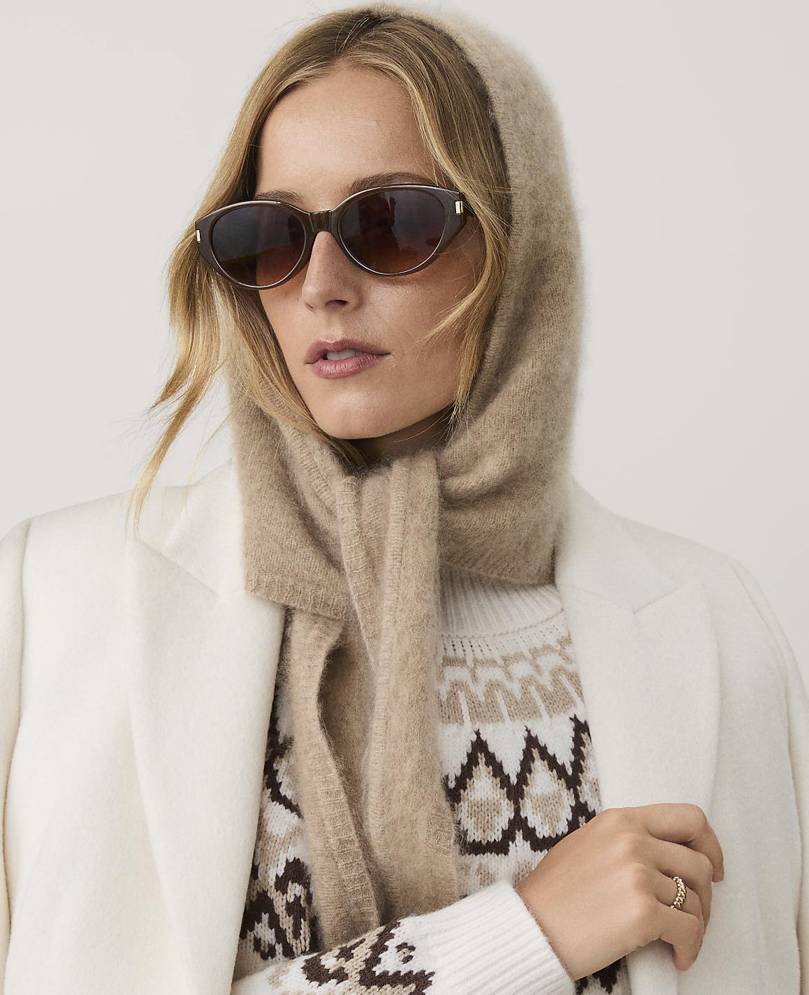 A woman wearing sunglasses, a beige hood-scarf, and a patterned sweater under a white coat poses against a plain light background.
