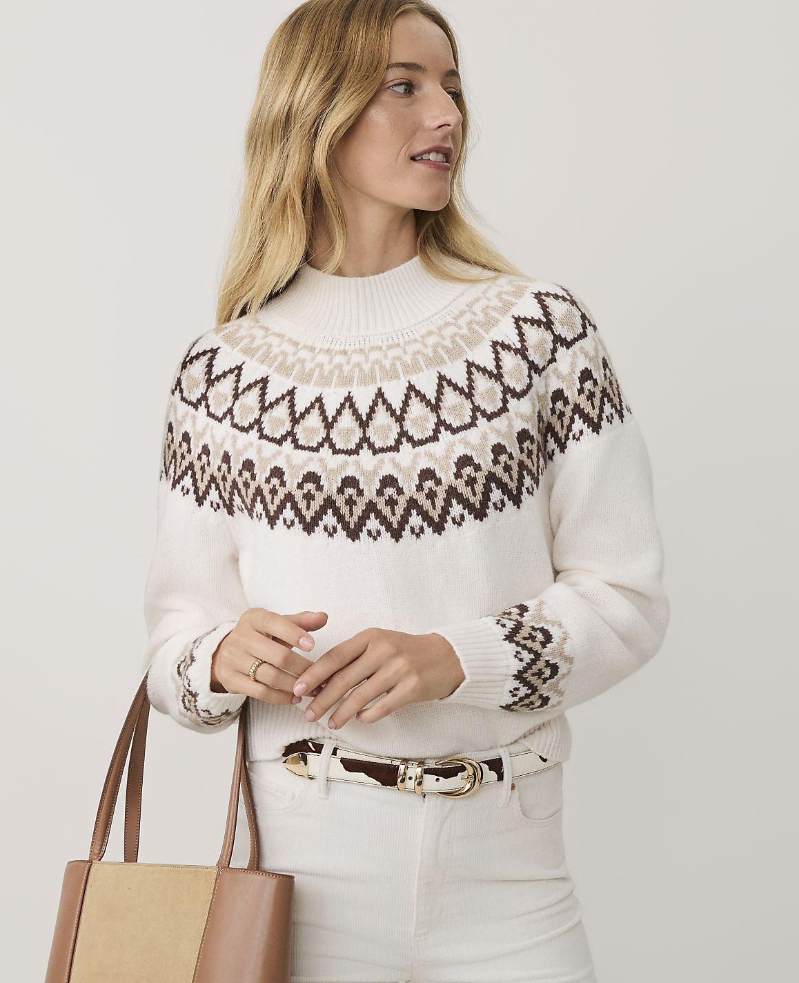 A blonde woman wearing a patterned white sweater and white pants stands against a plain light gray background.