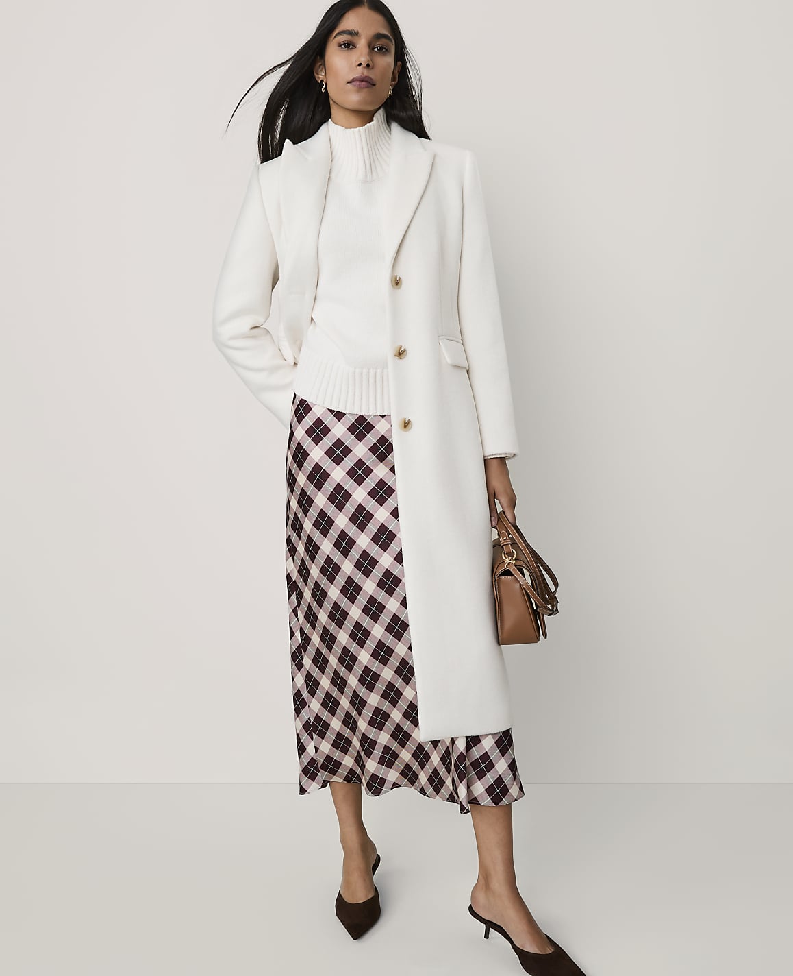 A woman wearing a white coat, white turtleneck, and a plaid skirt stands against a plain white background.
