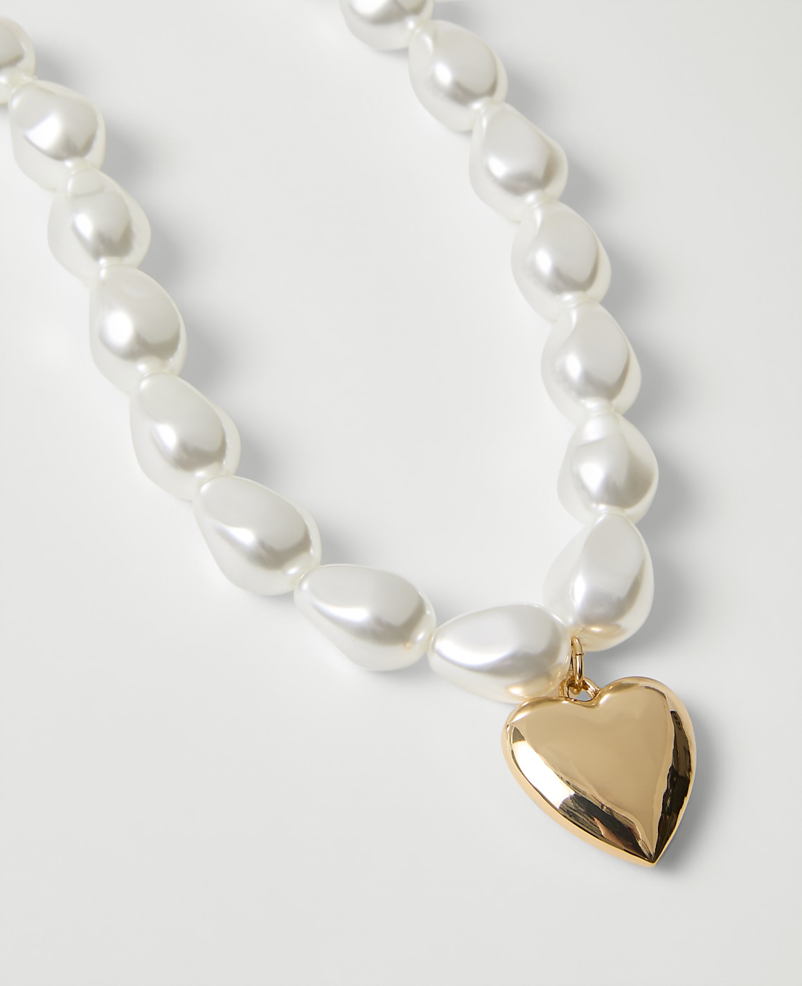 A necklace of large, iridescent white pearls with a gold heart-shaped pendant rests on a plain white background.