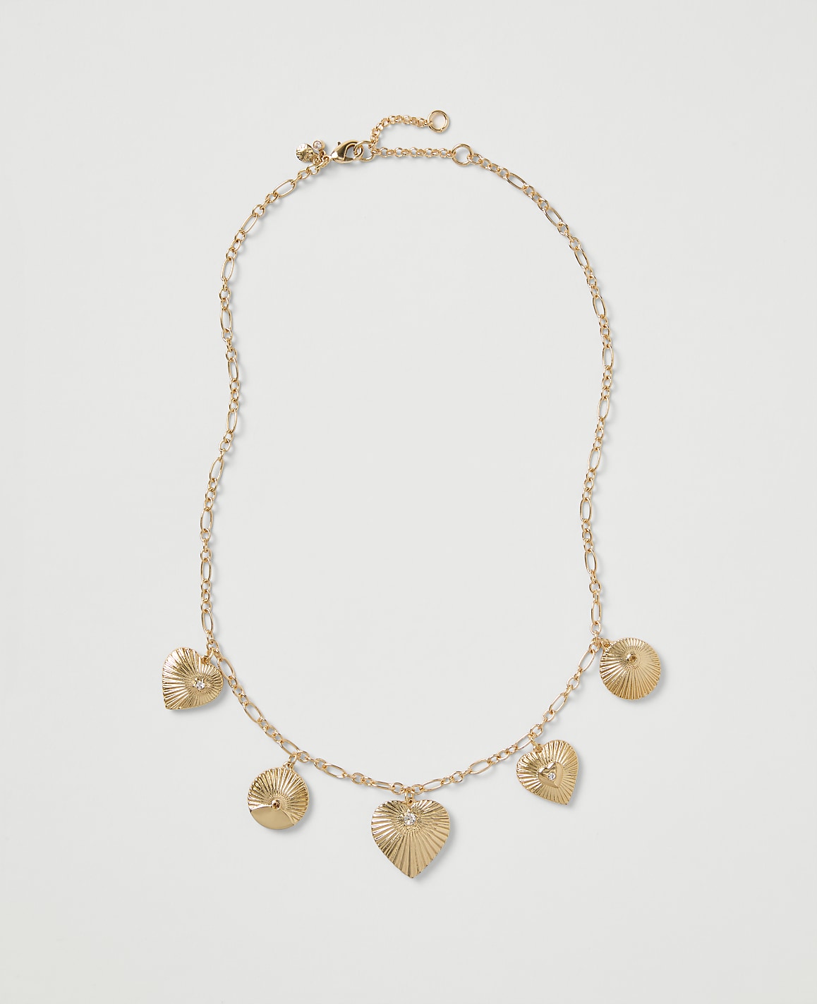 A gold chain necklace with heart and circular ribbed charms rests on a plain light gray background.