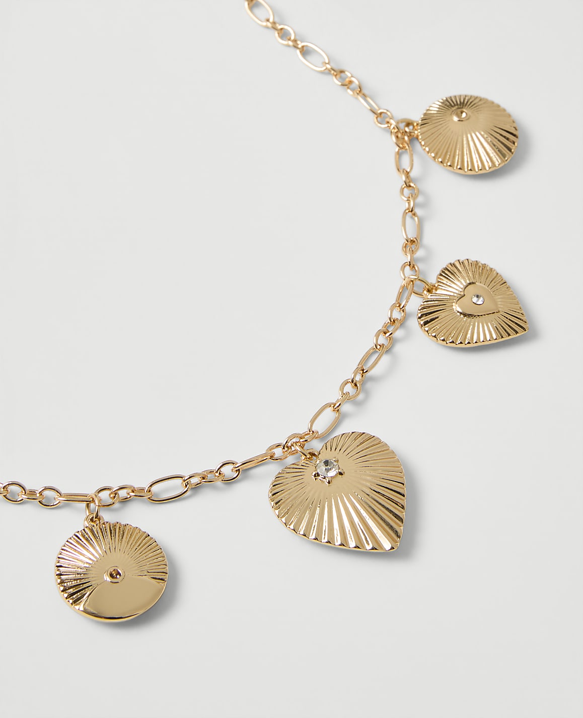 A gold-toned chain necklace with heart-shaped and circular ribbed charms rests on a plain white background.