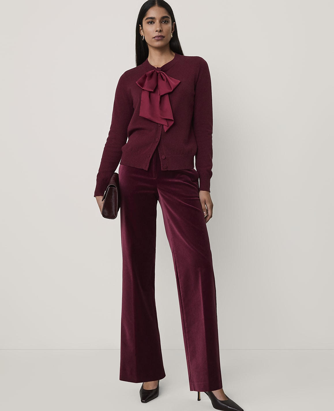 A woman wearing a burgundy bow-accented cardigan and matching velvet wide-leg trousers stands against a plain white background.