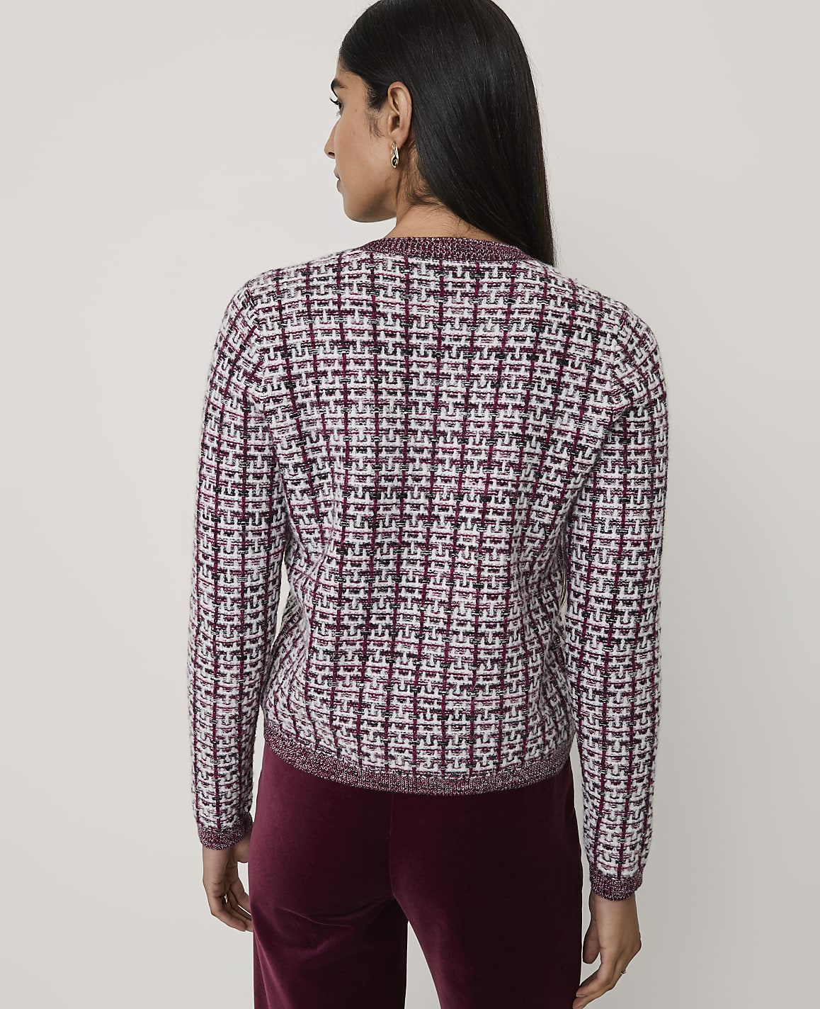 A woman stands with her back to the camera wearing a textured maroon and white tweed jacket and maroon velvet trousers against a plain light grey background.