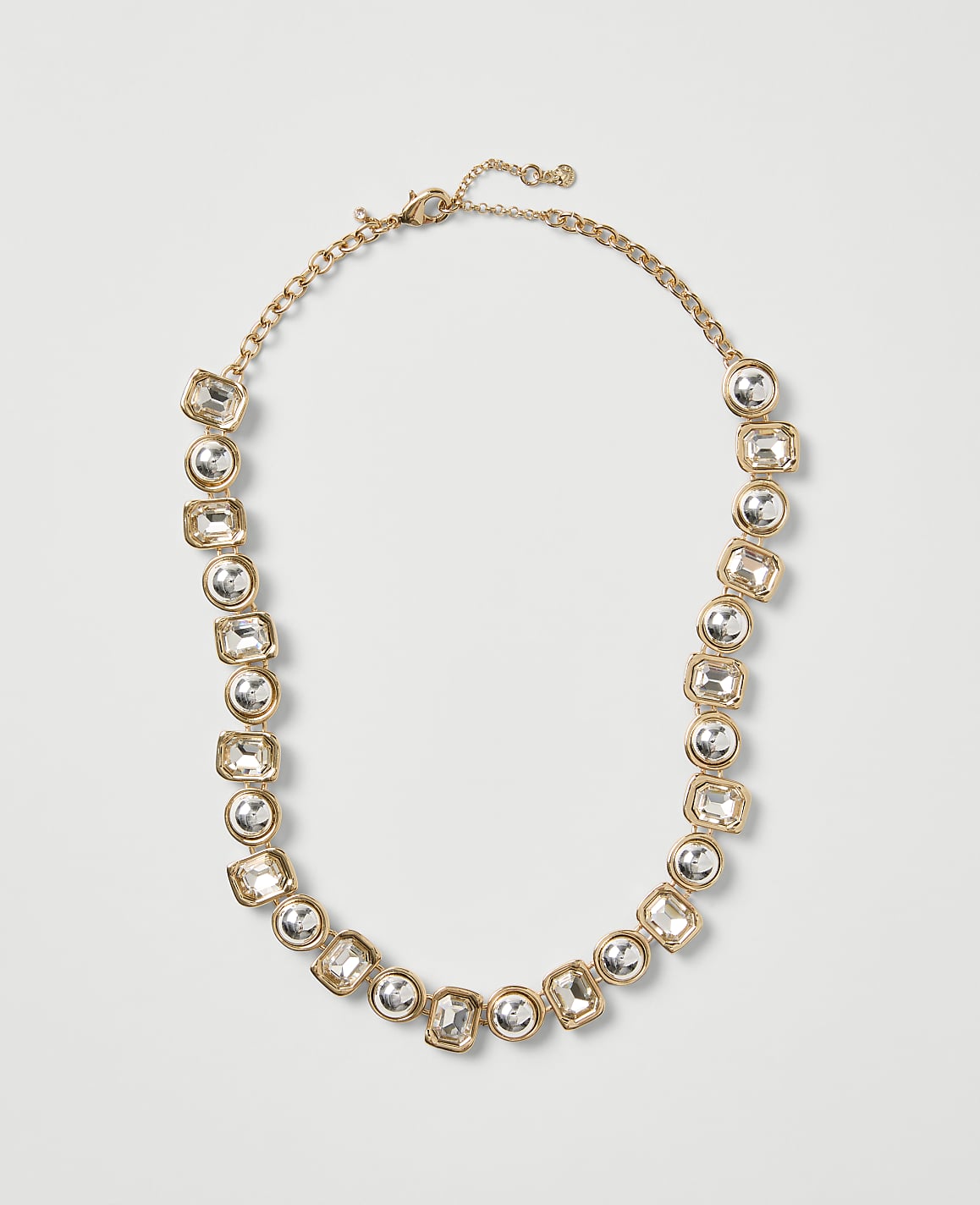 A gold-toned necklace with clear crystal stones is centered against a plain light gray background.