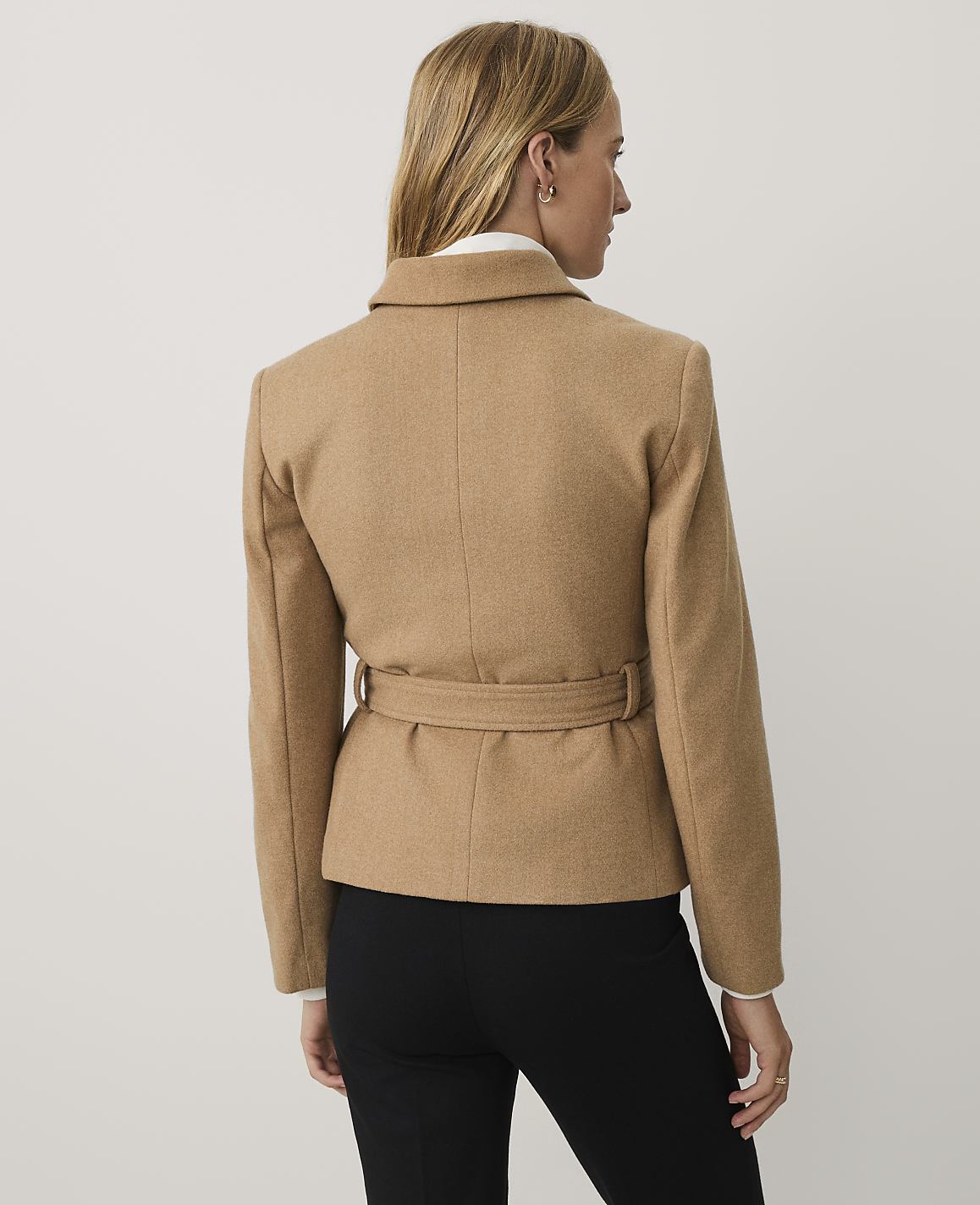 A woman stands with her back to the camera wearing a belted tan jacket and black trousers against a plain white background.