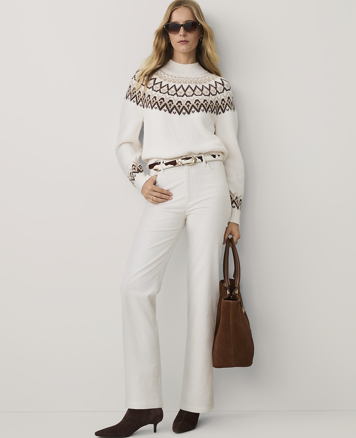 A woman wearing a patterned white sweater, white trousers, and brown heels stands against a plain white background.
