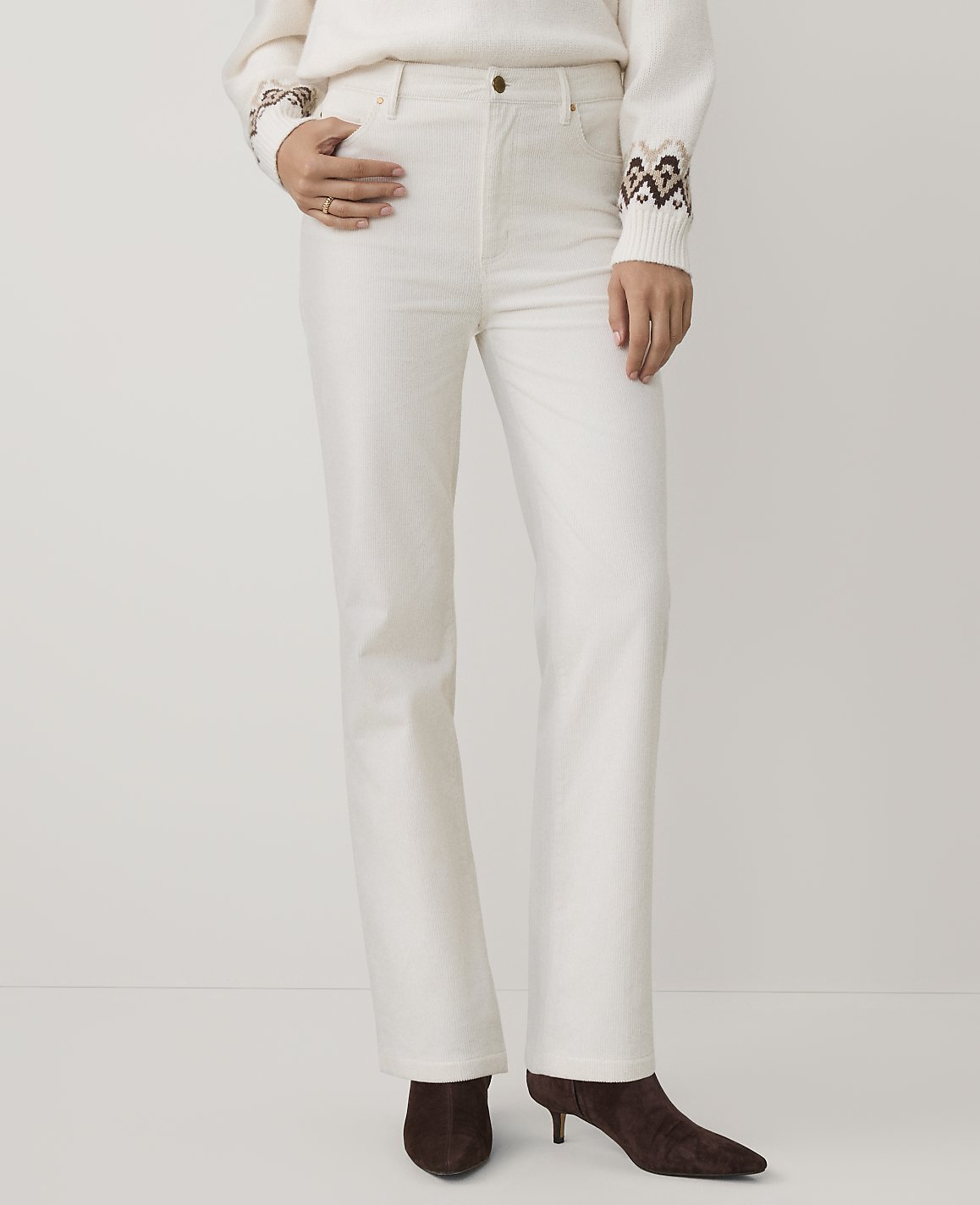 A person wearing a white sweater and white trousers with brown boots stands against a plain white background.