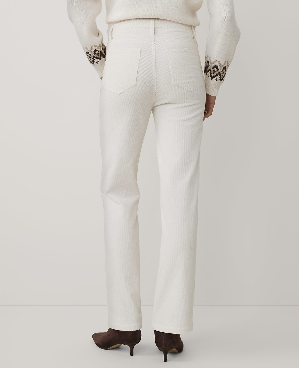 A person wearing a white sweater and white trousers with brown heels stands against a plain white background.