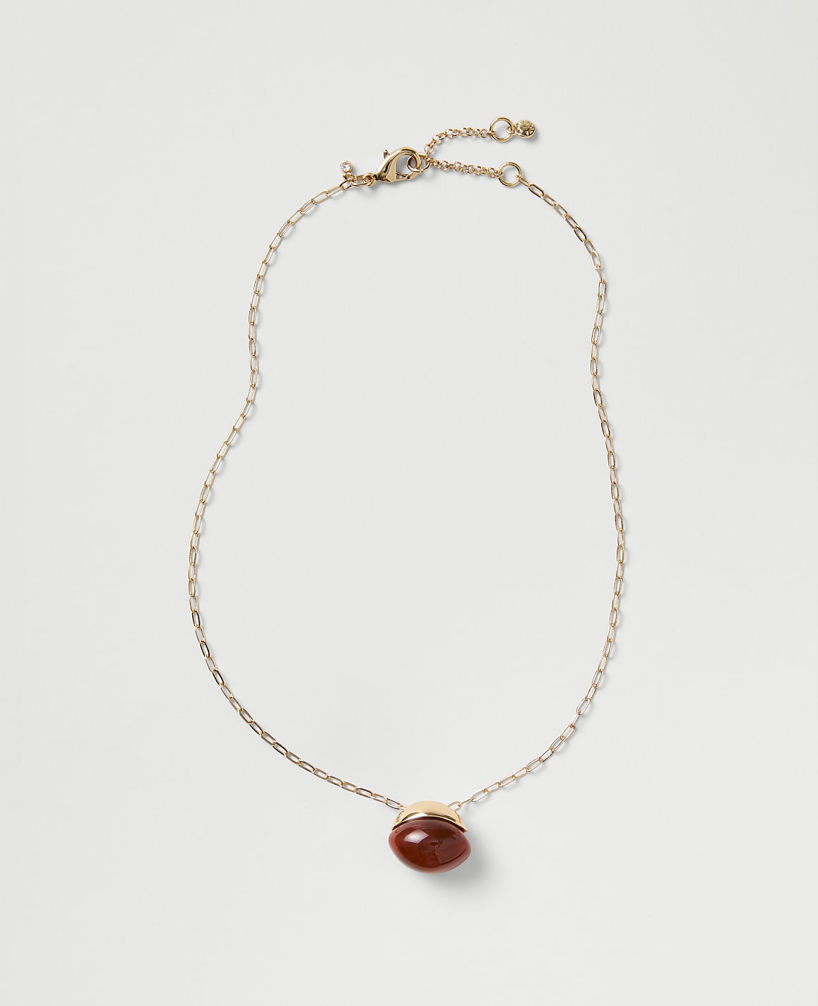 A gold chain necklace with a red oval gemstone pendant rests against a plain light gray background.