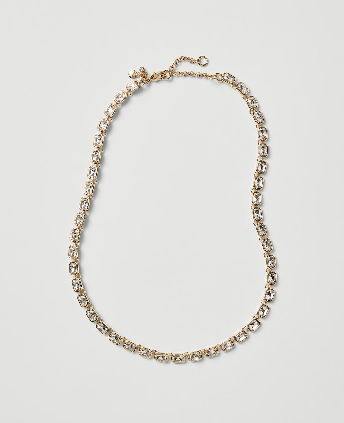 A gold-toned necklace with clear crystals is laid out against a plain, light gray background.