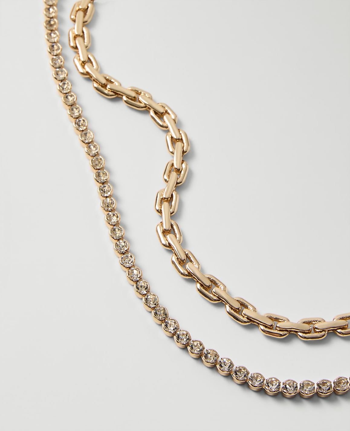 Two gold chains, one chunky and one embellished with crystals, lie draped across a plain light gray background.