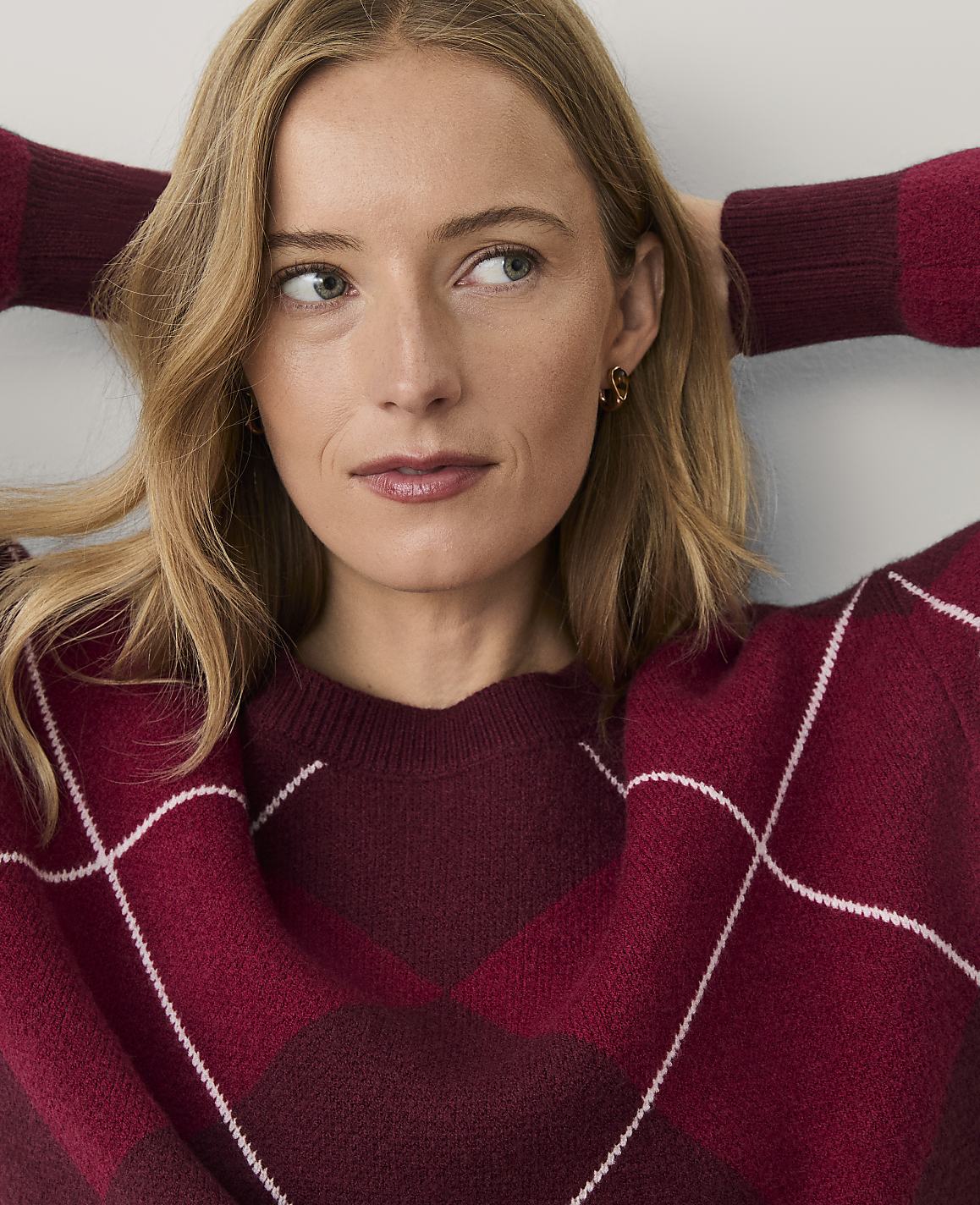 A blonde woman wearing a burgundy argyle sweater poses with her hands behind her head against a plain white background.