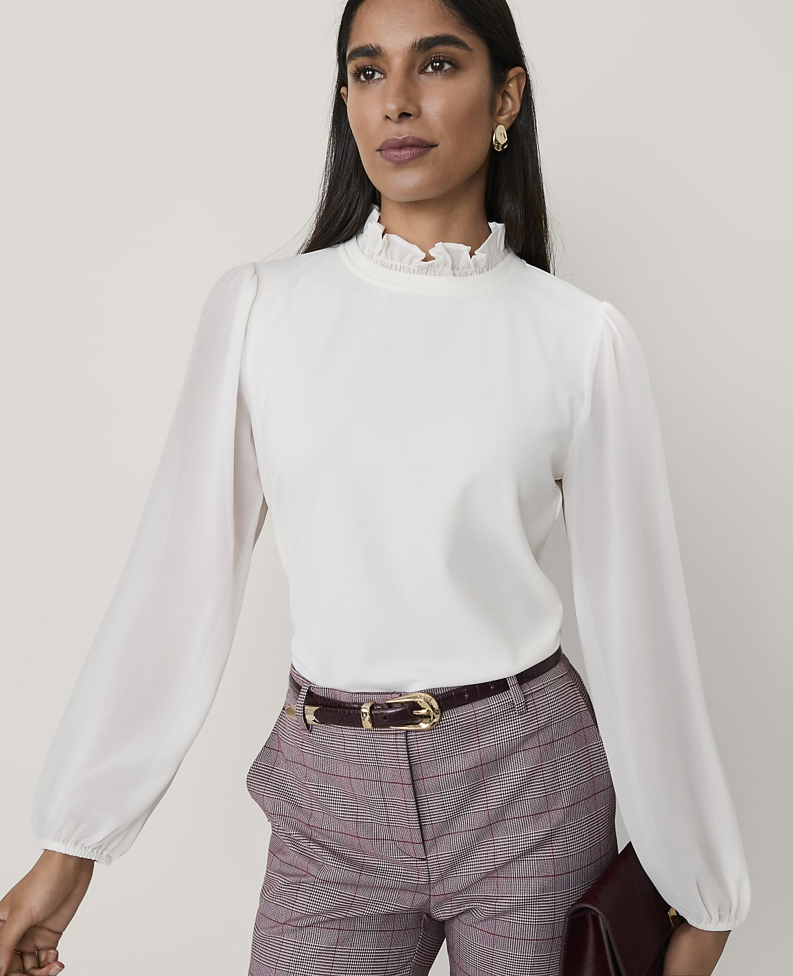A woman wearing a white ruffled blouse and plaid trousers stands against a plain, light-colored background.