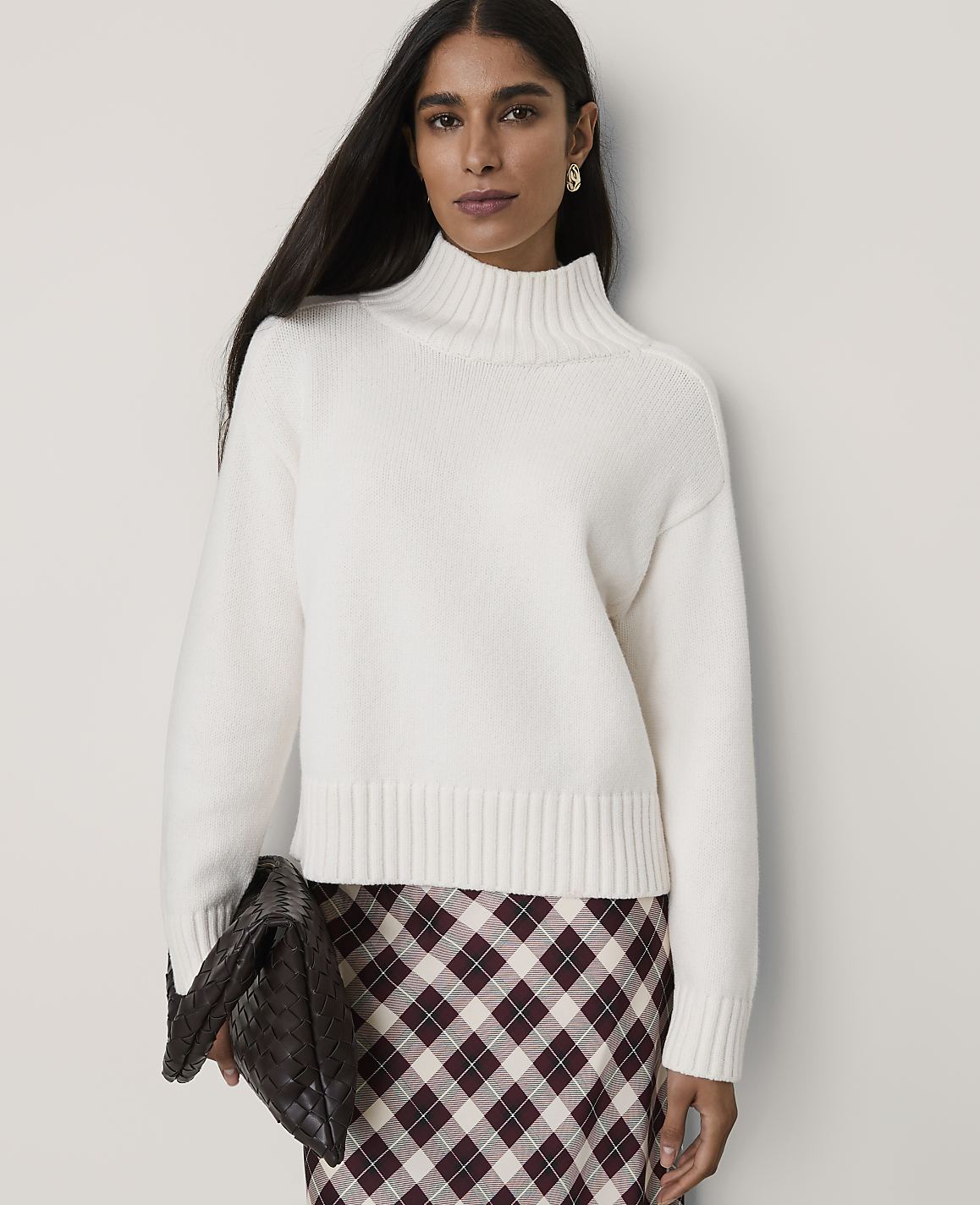A woman wearing a white mock-neck sweater and a plaid skirt stands against a plain, light-colored wall.