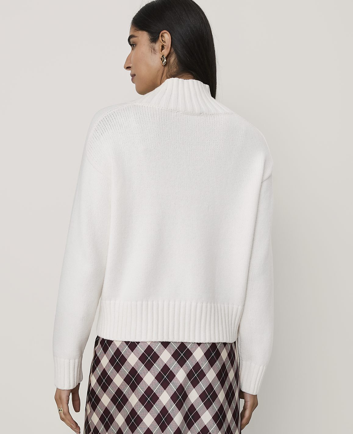 A woman wearing a white turtleneck sweater and a plaid skirt stands against a plain, light-colored background.