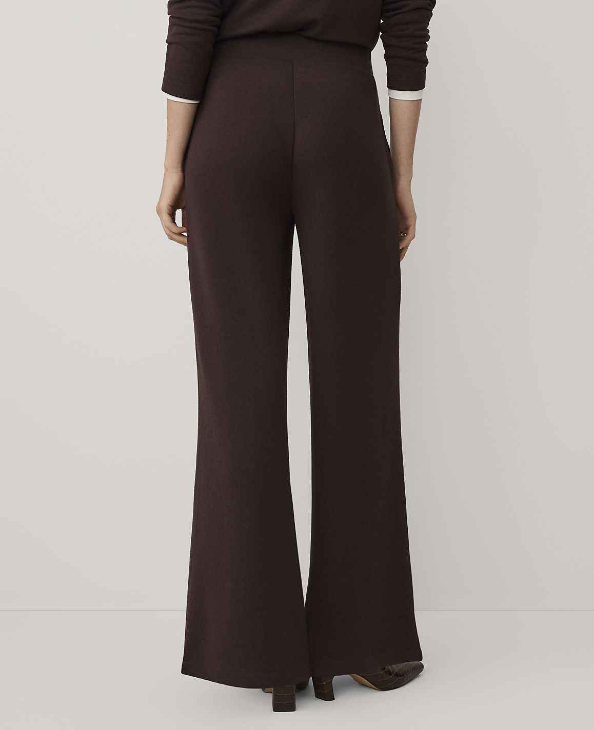 A person stands facing away from the camera wearing dark brown wide-leg trousers and a matching top against a plain, light-colored background.
