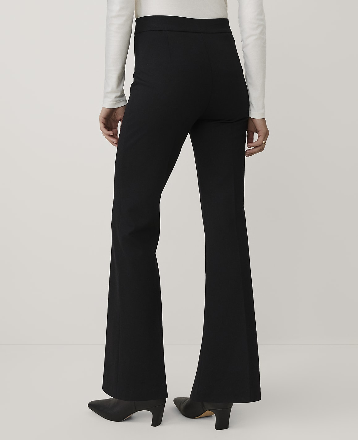 A person wearing a white long-sleeved top, black flared trousers, and black heels stands against a plain light gray background.