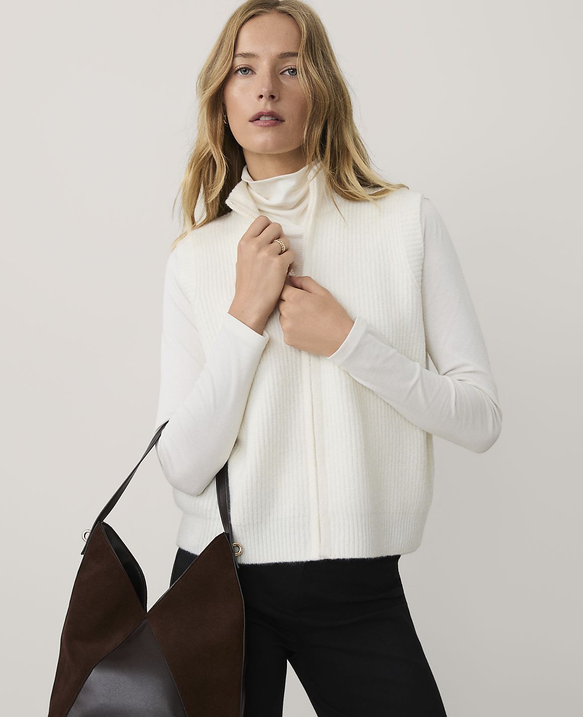 A blonde woman wearing a white turtleneck and vest stands against a plain, light-colored background while holding a large brown handbag.
