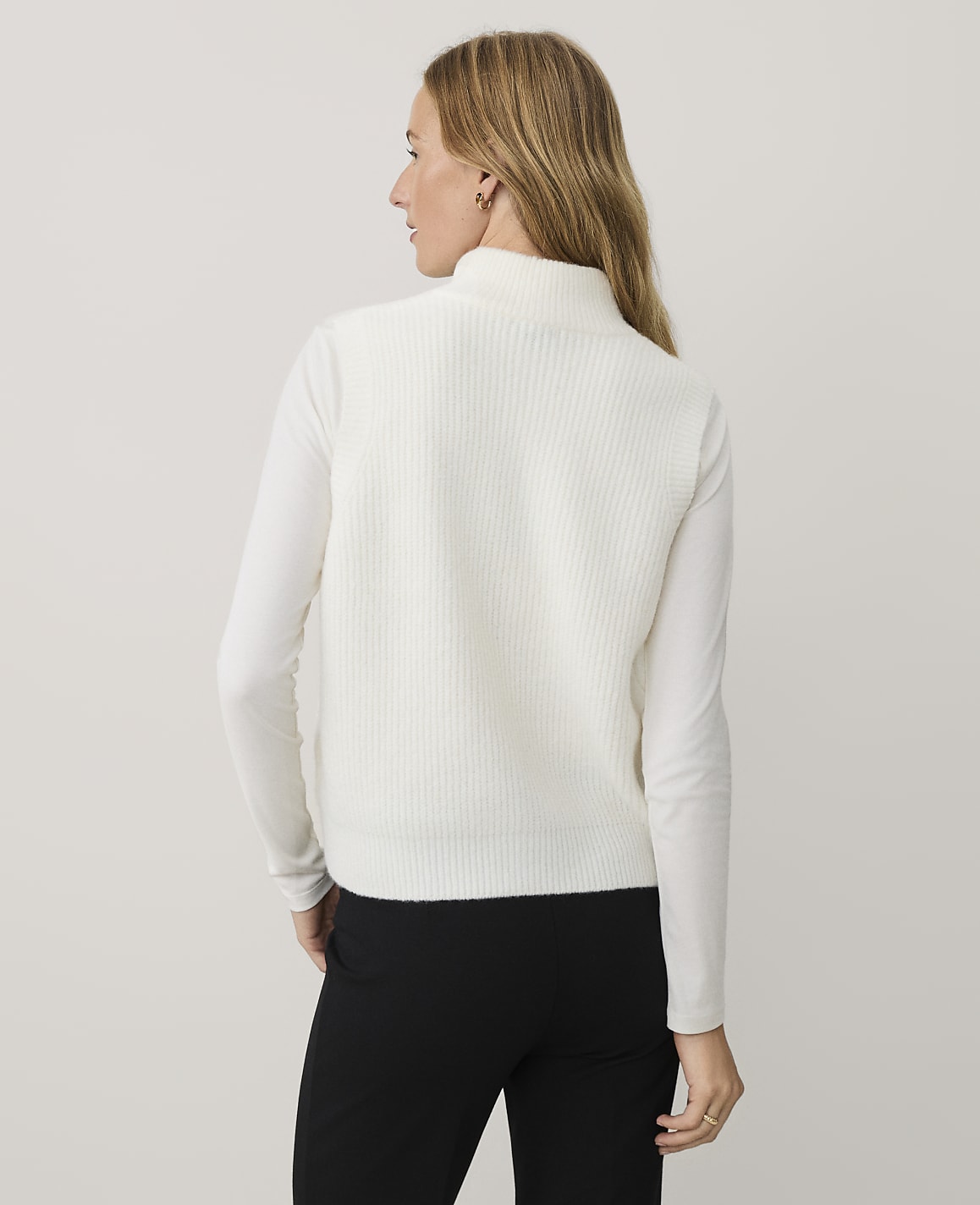 A woman wearing a white ribbed sweater and black pants stands against a plain, light-colored background.