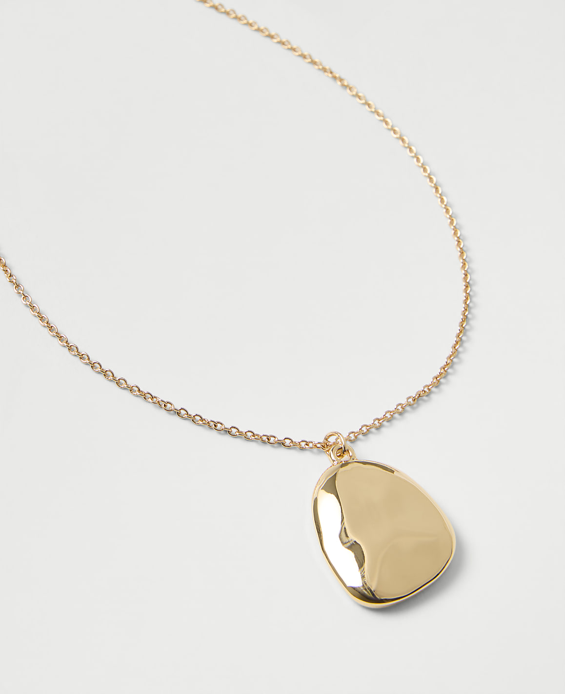 A gold chain necklace with a polished, organic-shaped pendant rests on a plain light gray background.