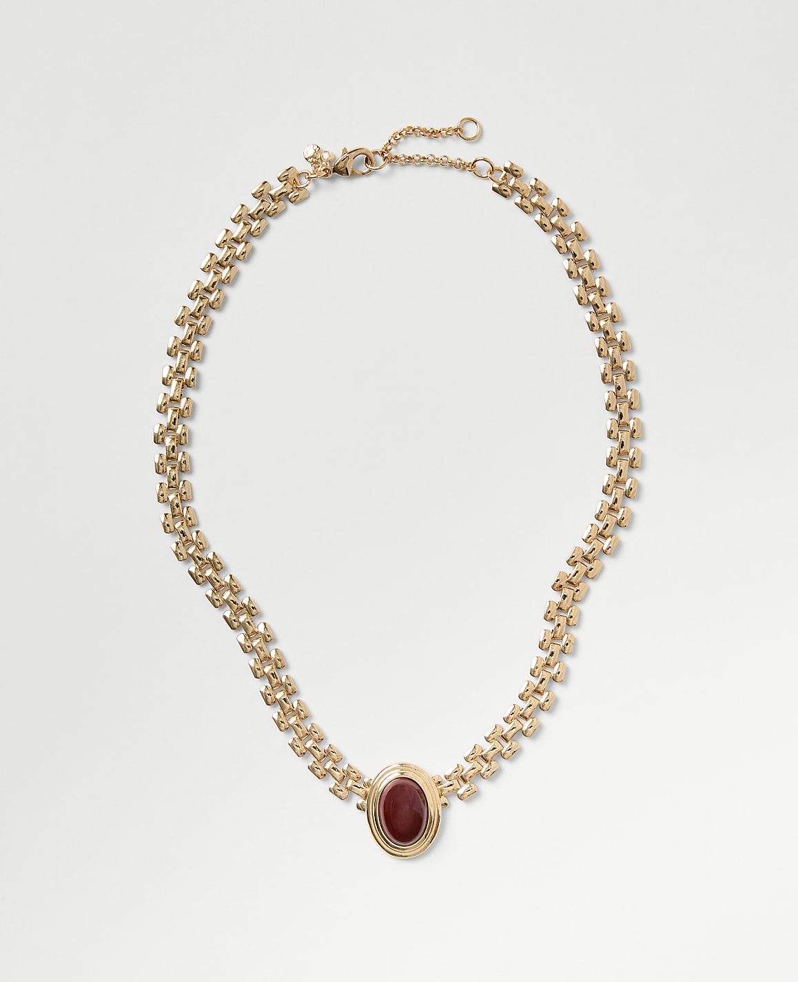 A gold chain necklace with a red oval gemstone pendant rests on a plain light gray background.