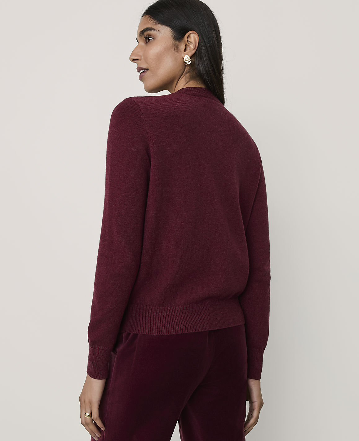A woman wearing a burgundy sweater and matching velvet trousers stands against a plain, light-colored background.