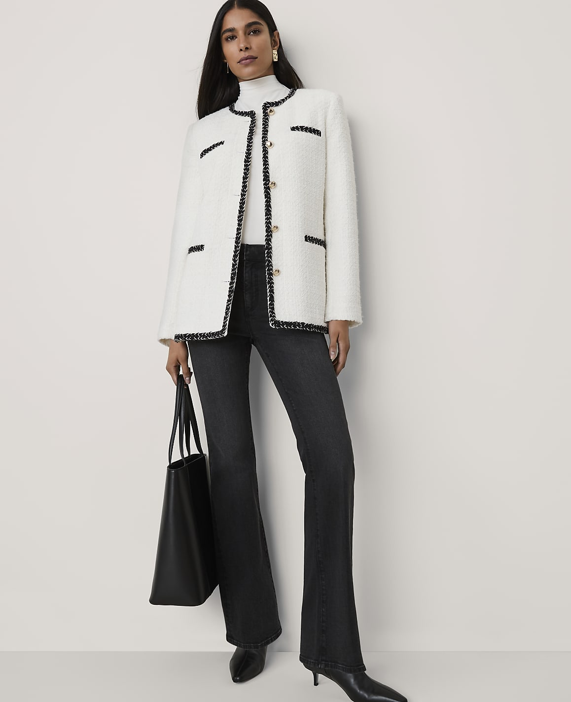 A woman wearing a white tweed jacket, black flared jeans, and holding a black tote bag stands against a plain light gray background.