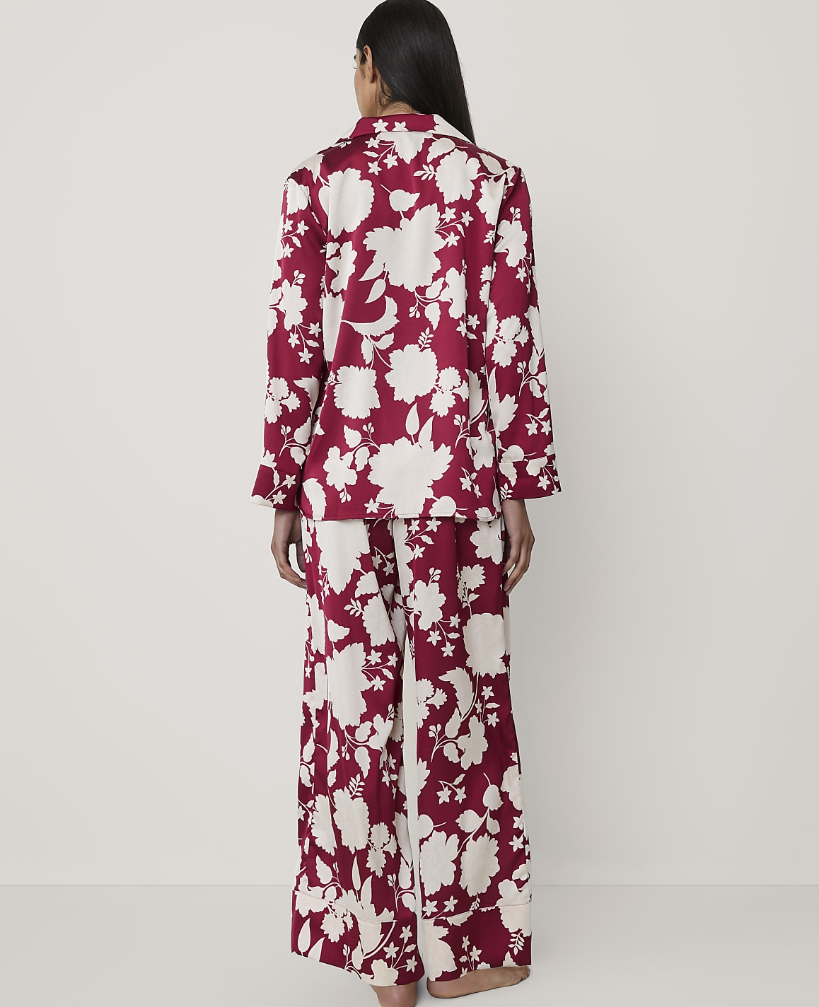 A woman stands with her back to the camera wearing red and white floral pajamas against a plain, light-colored background.