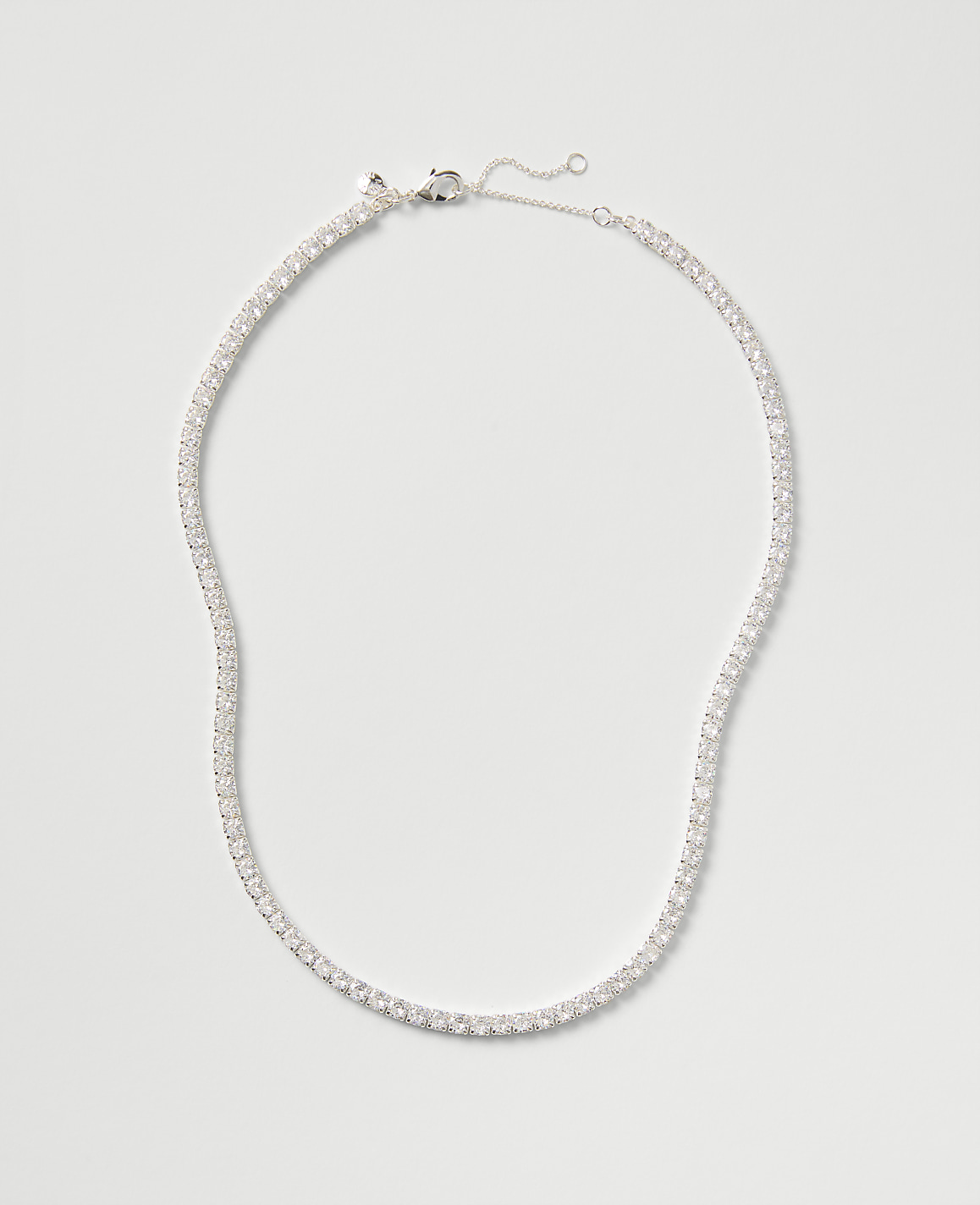 A sparkling tennis necklace with clear stones rests against a plain, light gray background.