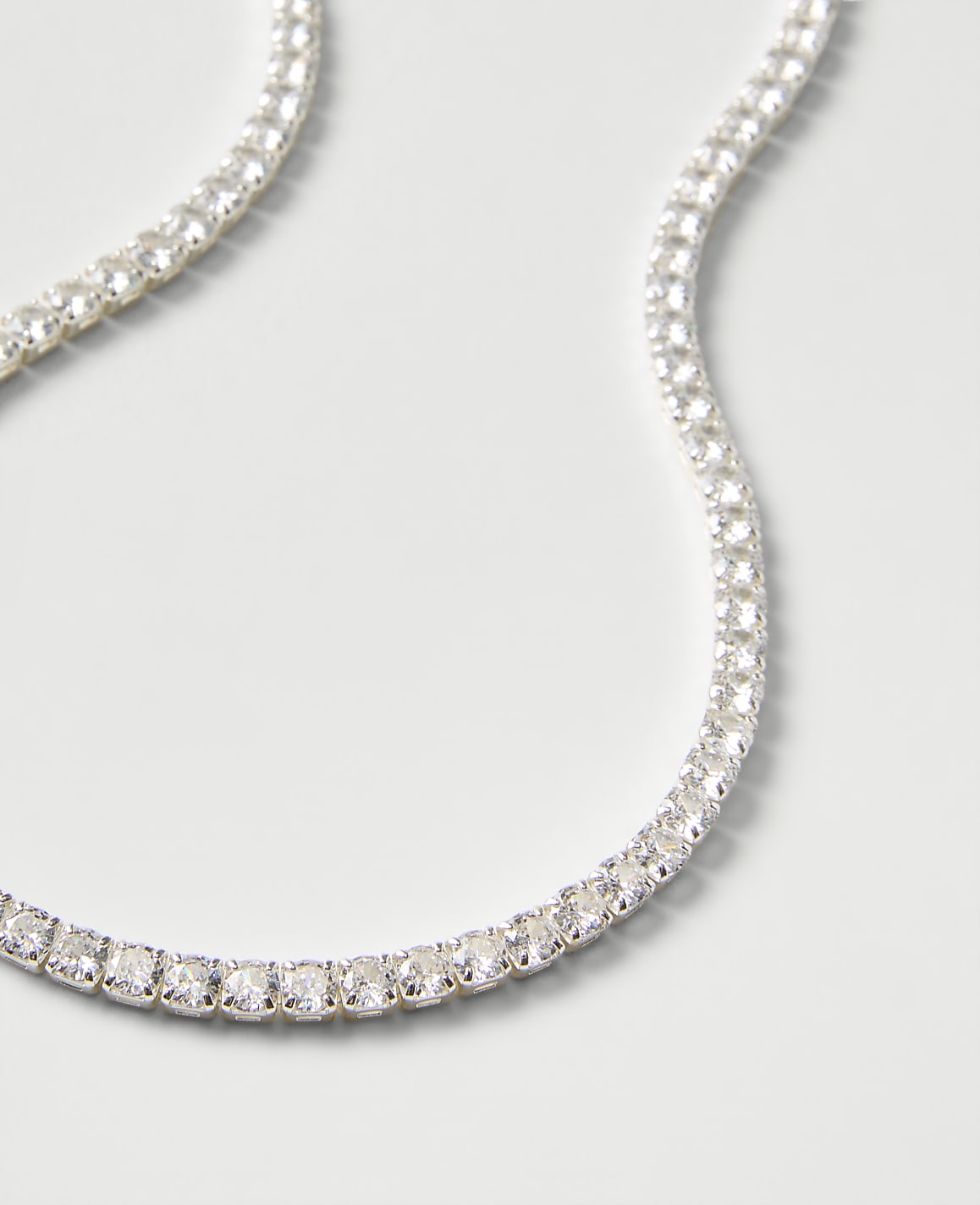 A sparkling tennis necklace of clear gemstones curves across a plain, light gray background.