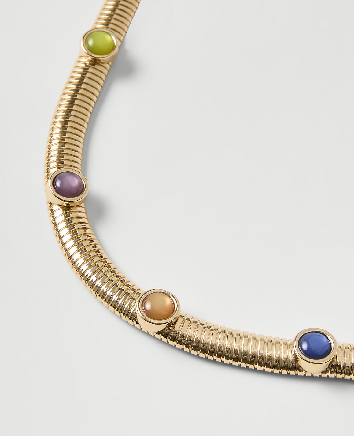 A gold ribbed necklace featuring colorful gemstones is set against a plain light gray background.