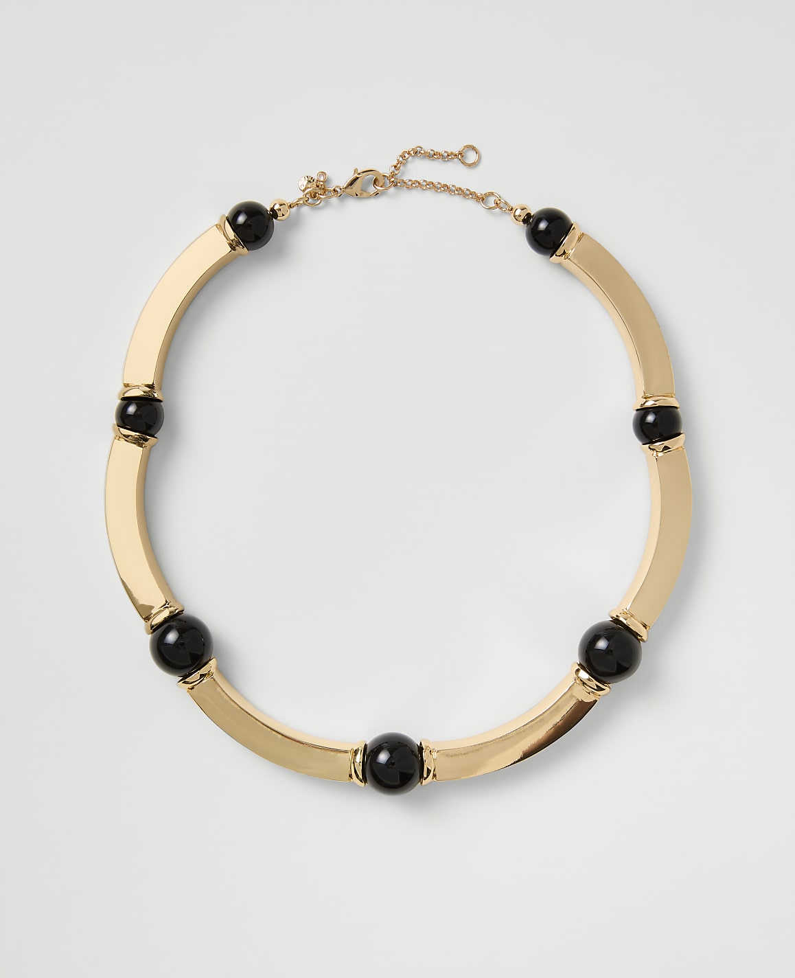A gold-toned necklace with black spherical beads rests on a plain light gray background.