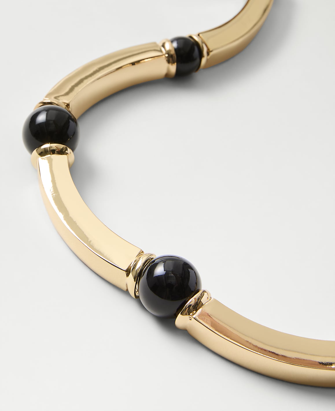 A gold-toned curved necklace with black spherical beads rests on a plain white background.