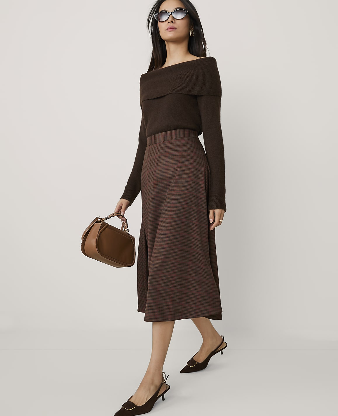 A woman wearing a brown off-the-shoulder top, a plaid skirt, and sunglasses stands against a plain white background.