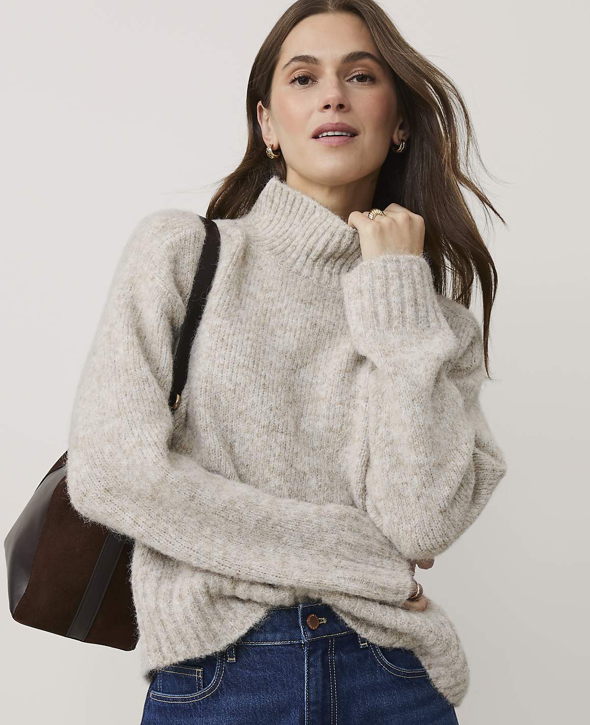 A woman wearing a beige turtleneck sweater and blue jeans stands against a plain, light-colored background.