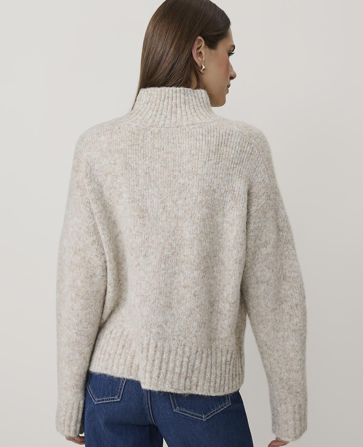 A woman wearing a beige knit sweater and blue jeans stands against a plain, light-colored background.