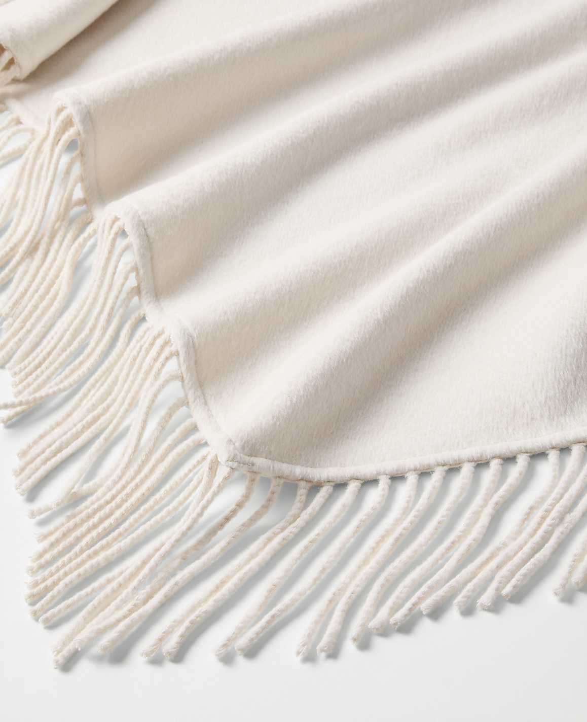 A cream-colored fringed blanket is draped in soft folds over a plain white background.