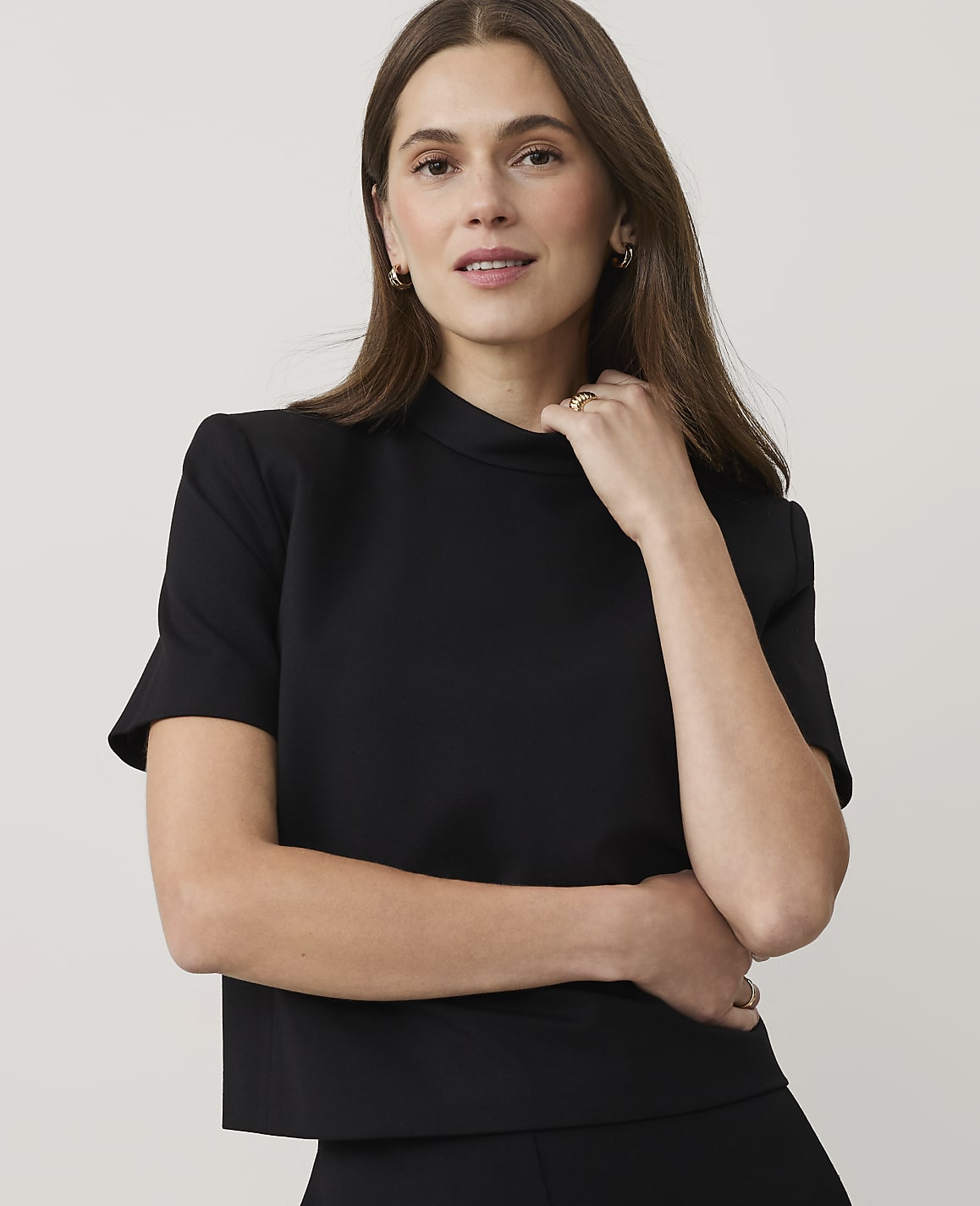 A woman wearing a black short-sleeved top and gold jewelry poses against a plain, light-colored background.