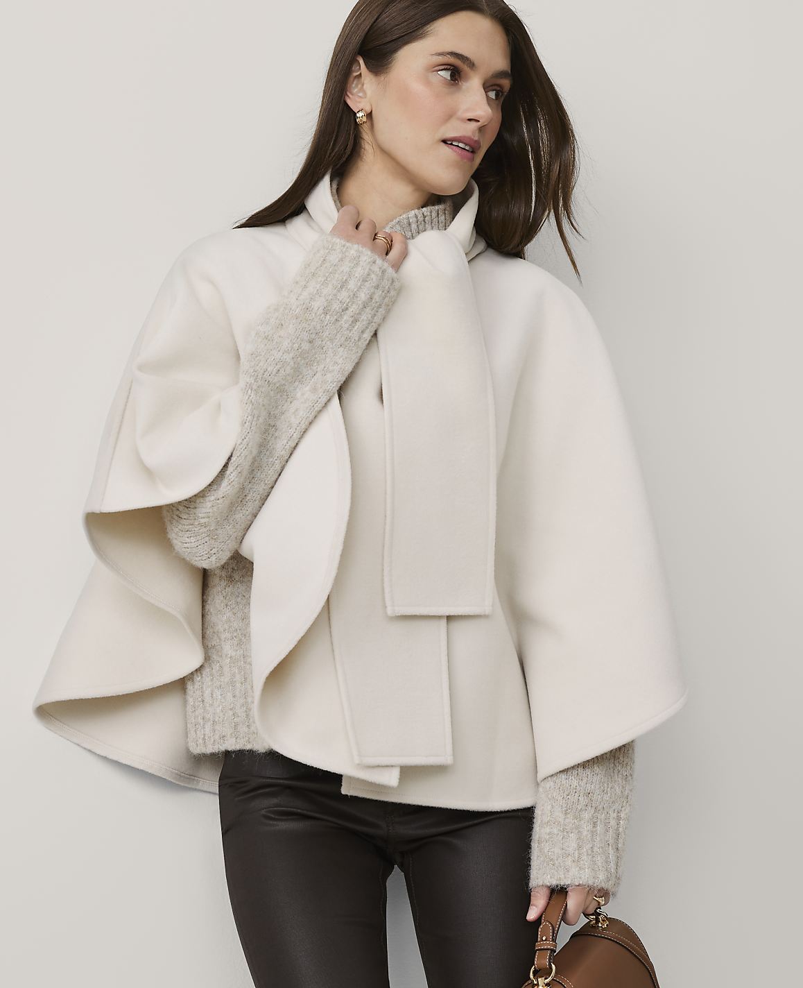 A woman wearing a cream-colored cape and brown leather pants stands against a plain, light-grey background.