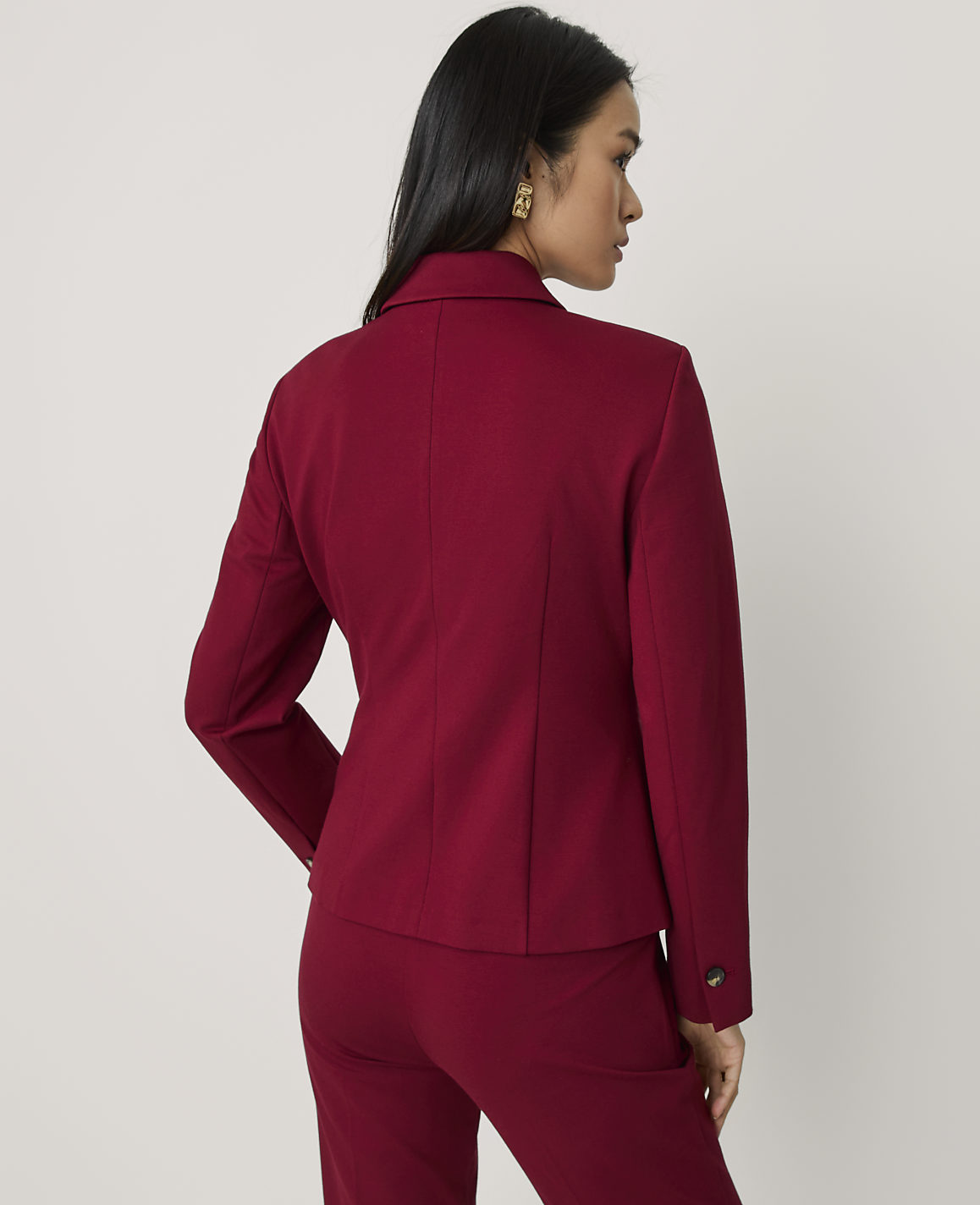 A woman wearing a red blazer and matching trousers stands with her back to the camera against a plain light grey background.