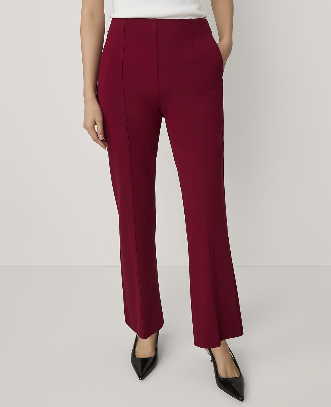 A person wears high-waisted burgundy trousers and black pointed heels against a plain, light-colored background.