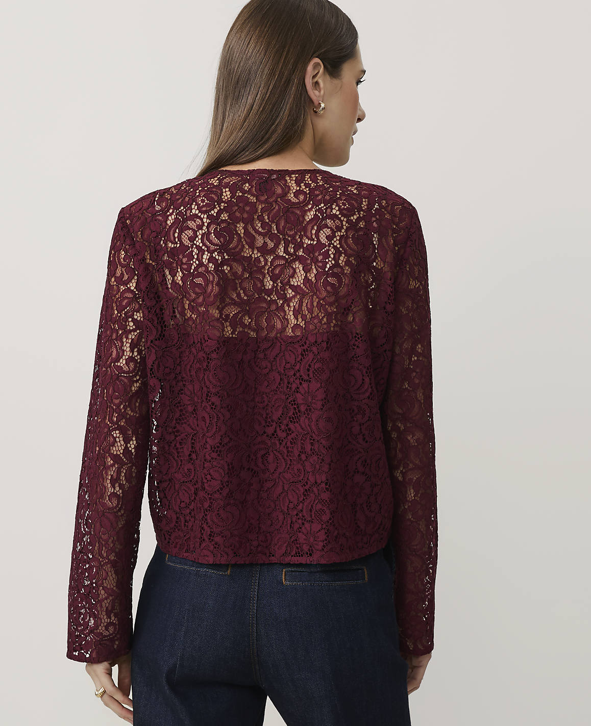 A woman wearing a burgundy lace top and dark blue jeans stands against a plain white background.