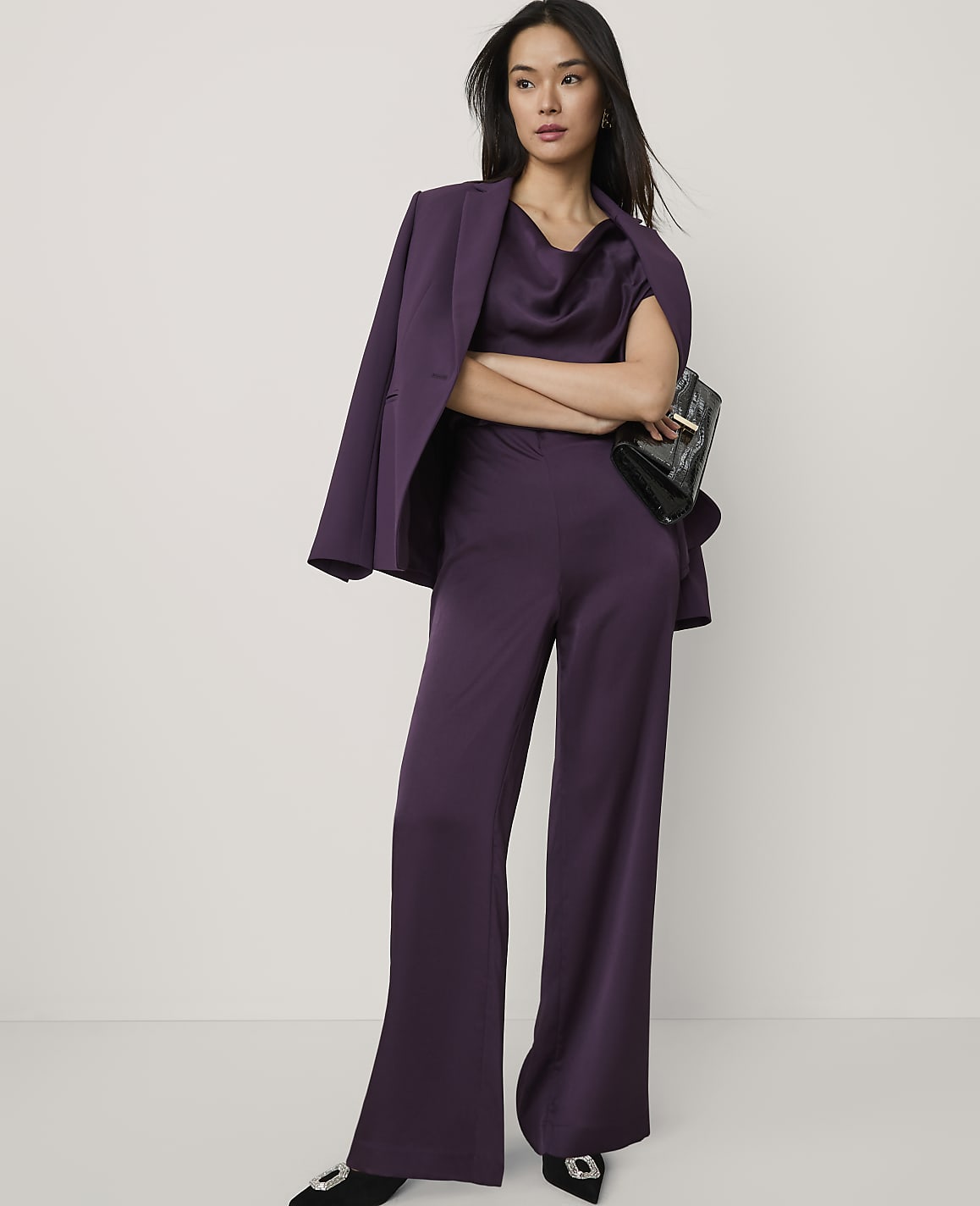A woman wearing a purple satin suit and holding a black clutch stands against a plain white background.